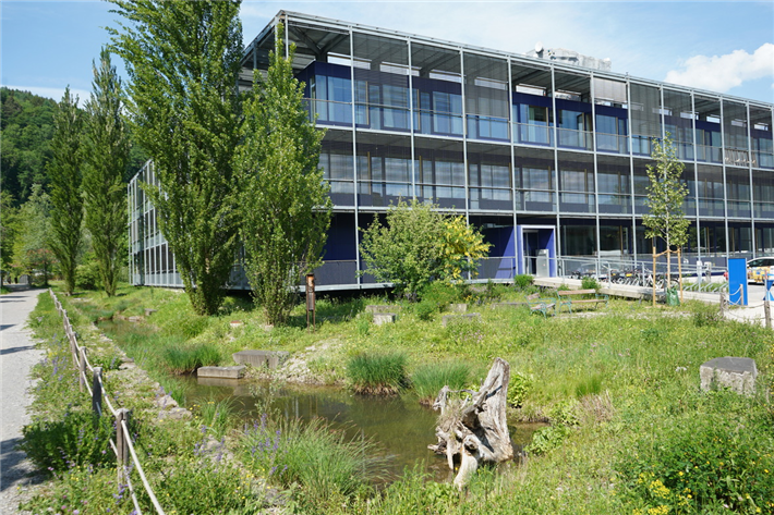 Hitzeresilienz Gewerbegebiet: Das Gelände der Firma Omicron in Klaus in Vorarlberg zeigt, wie ein Unternehmen gemeinsam mit der Gemeinde von der Renaturierung eines Bachlaufs profitieren kann: Die Hochwassergefahr wurde reduziert, das Mikroklima vor Ort verbessert, die biologische Vielfalt bereichert und die Aufenthaltsqualität für die Mitarbeiter erhöht. © Bodensee-Stiftung